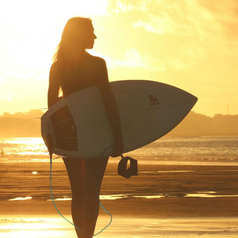 Eine Silhouette einer Frau, die ein Surfbrett hält und an einem Strand im Sonnenuntergang mit goldenem Himmel steht.